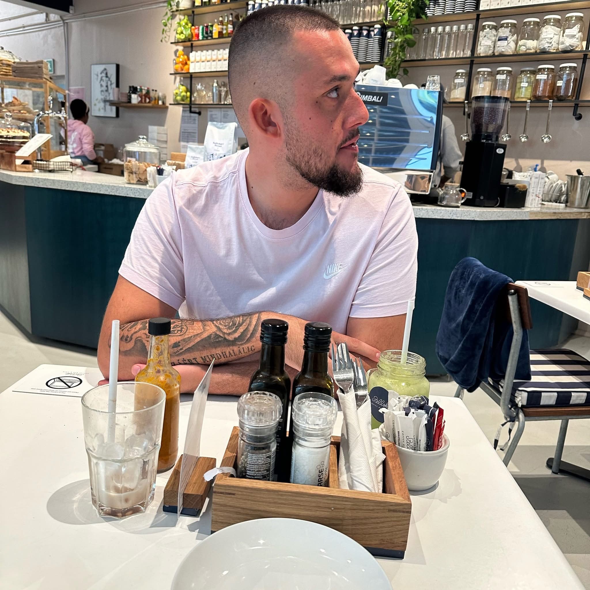 Ein tätowierter Mann im rosa T-Shirt sitzt an einem Cafétisch mit Getränken und Gewürzen.